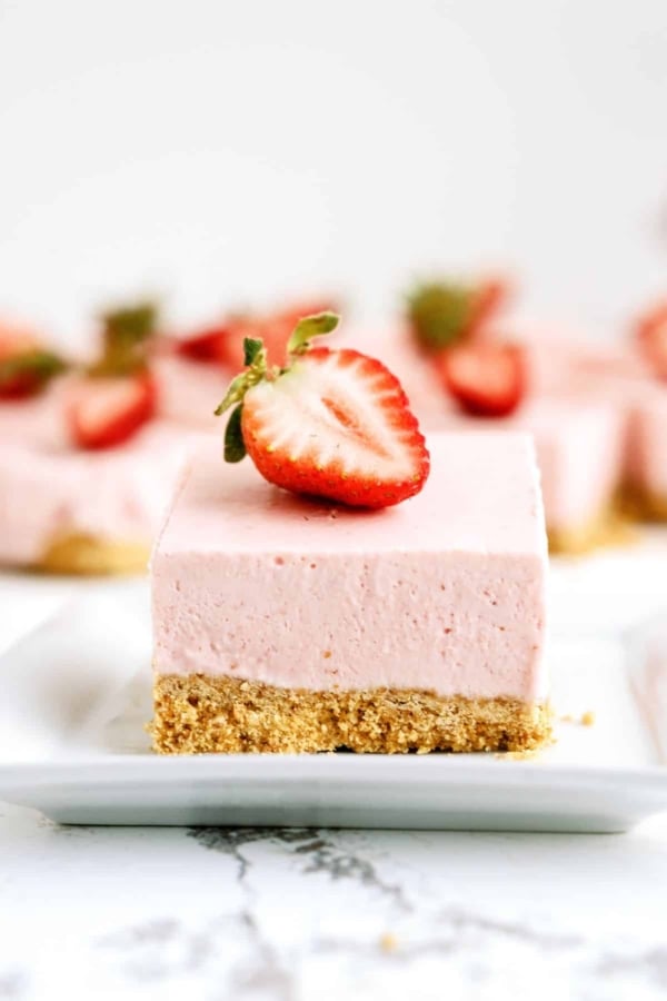 A No Bake Strawberry Cheesecake Bar on a plate with a slice of strawberry on top. More Cheesecake bars on plates in the background.