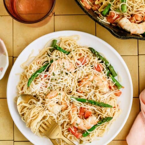 A plate of pasta with shrimp, asparagus, tomatoes, and grated cheese, served with a fork and knife, next to a glass of pink beverage and a skillet containing more pasta.