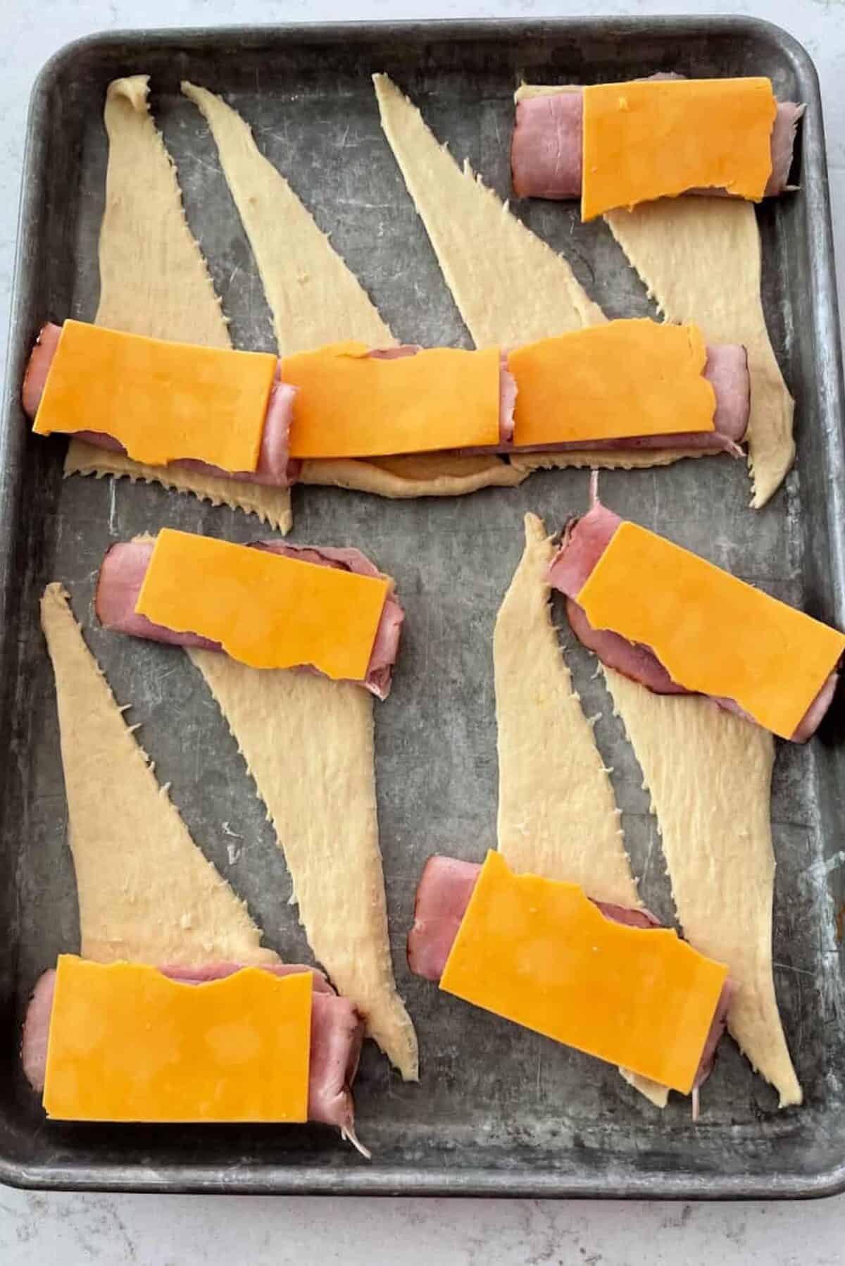 Unbaked crescent rolls on a baking sheet, each topped with a slice of ham and cheddar cheese.