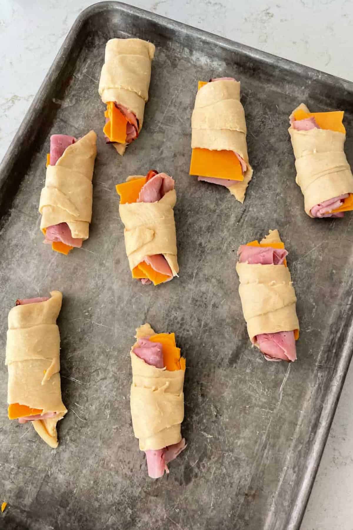 Unbaked crescent rolls filled with ham and cheese on a metal baking sheet.