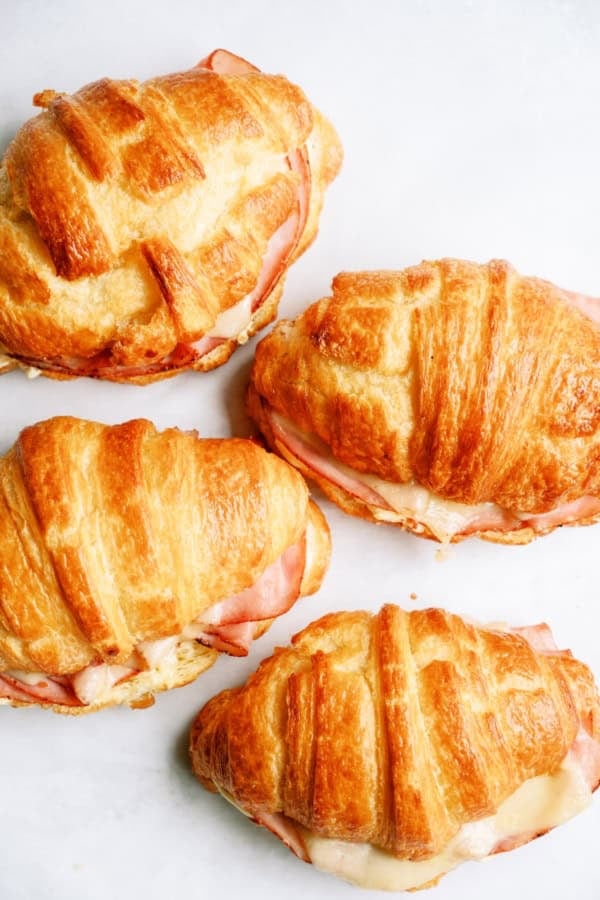 Four croissant sandwiches filled with ham and melted cheese are arranged on a white surface.