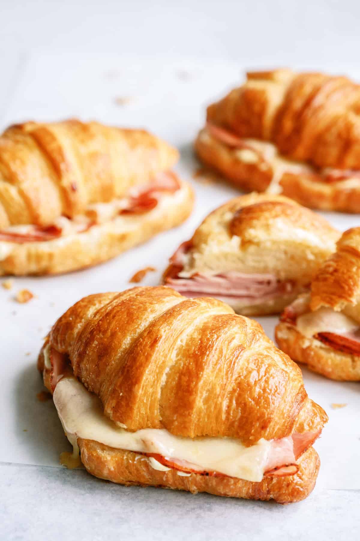 Four baked croissant sandwiches filled with ham and melted cheese are arranged on a white surface.