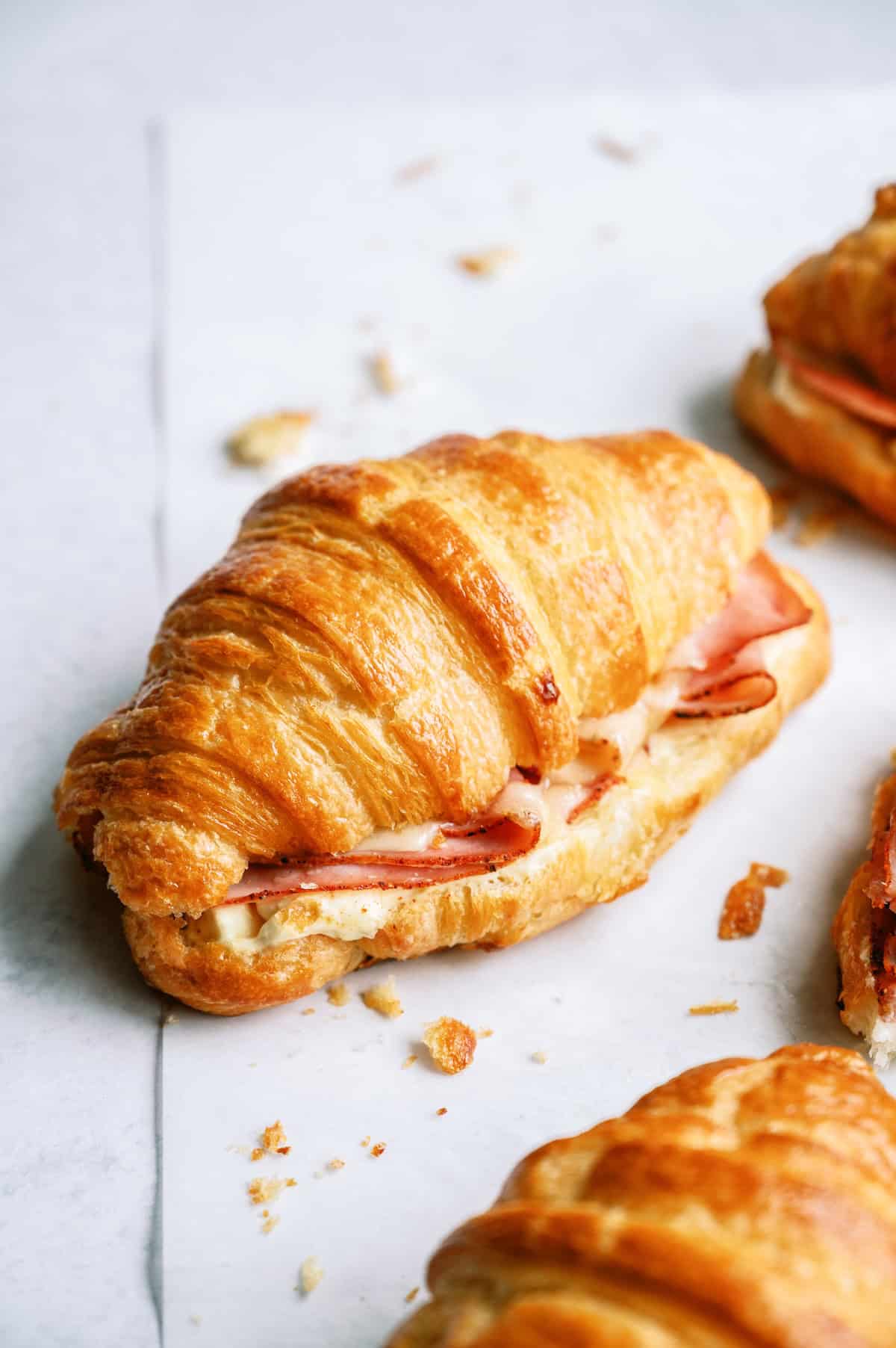 A croissant sandwich filled with ham and cheese sits on parchment paper, with crumbs scattered around.