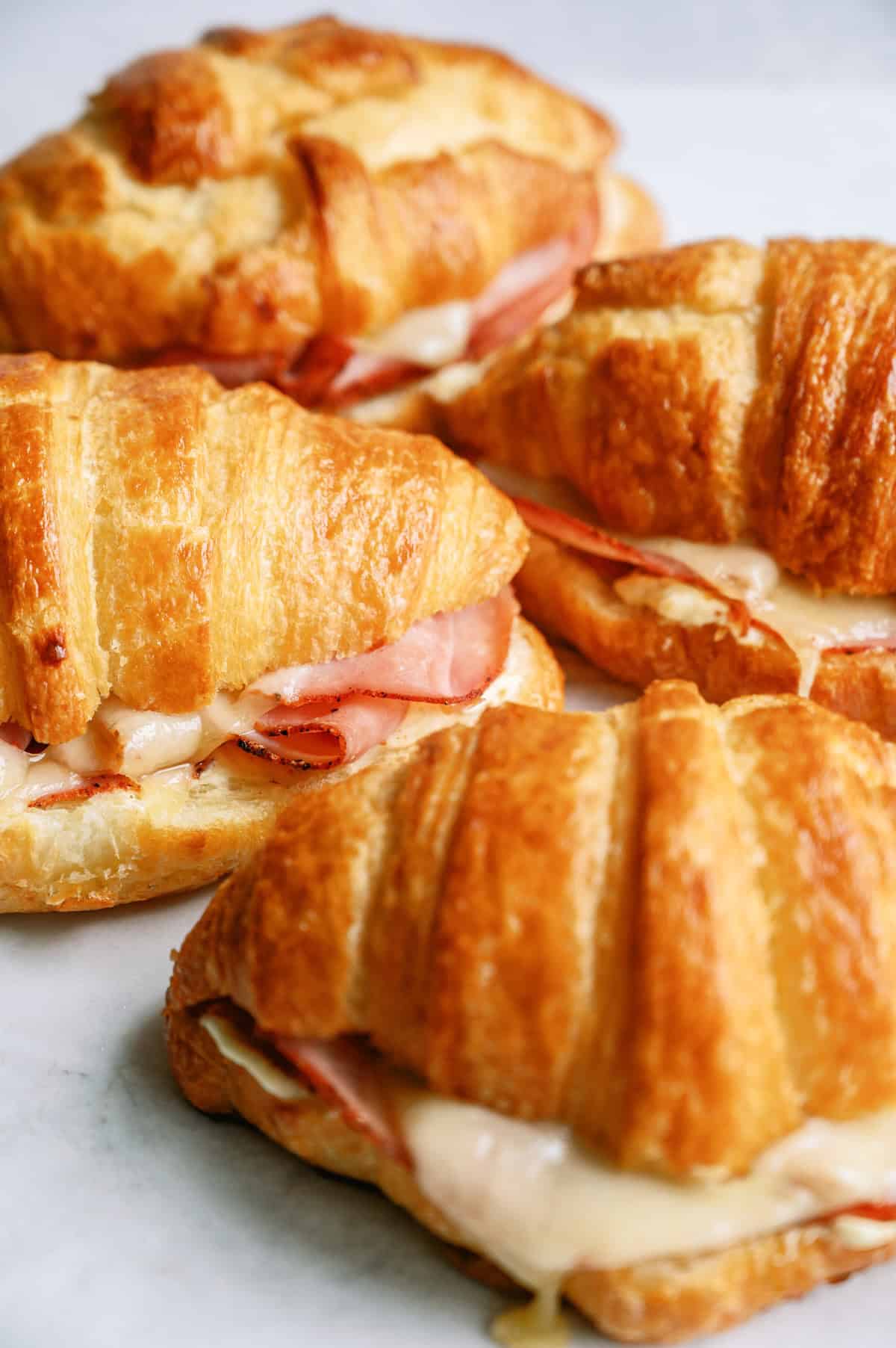 Four croissant sandwiches filled with ham and melted cheese are arranged on a white surface.