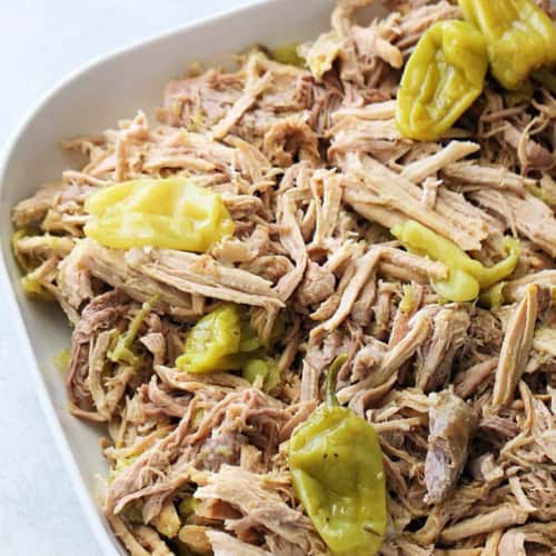Shredded cooked pork topped with several whole pepperoncini peppers served in a white dish.