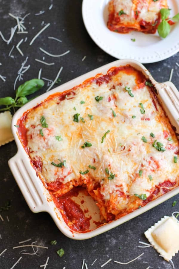 Freshly baked ravioli lasagna in a square ceramic baking dish, covered in melted bubbly cheese, marinara sauce, and chopped fresh basil, with one serving scooped out and plated in the background.