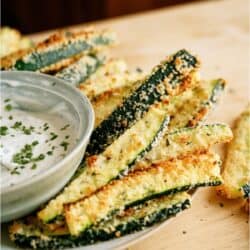 Plate of breaded zucchini sticks with a small bowl of dipping sauce, garnished with chopped herbs.