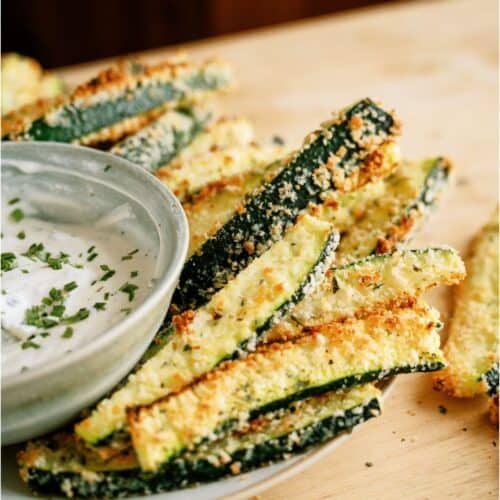 Plate of breaded zucchini sticks with a small bowl of dipping sauce, garnished with chopped herbs.