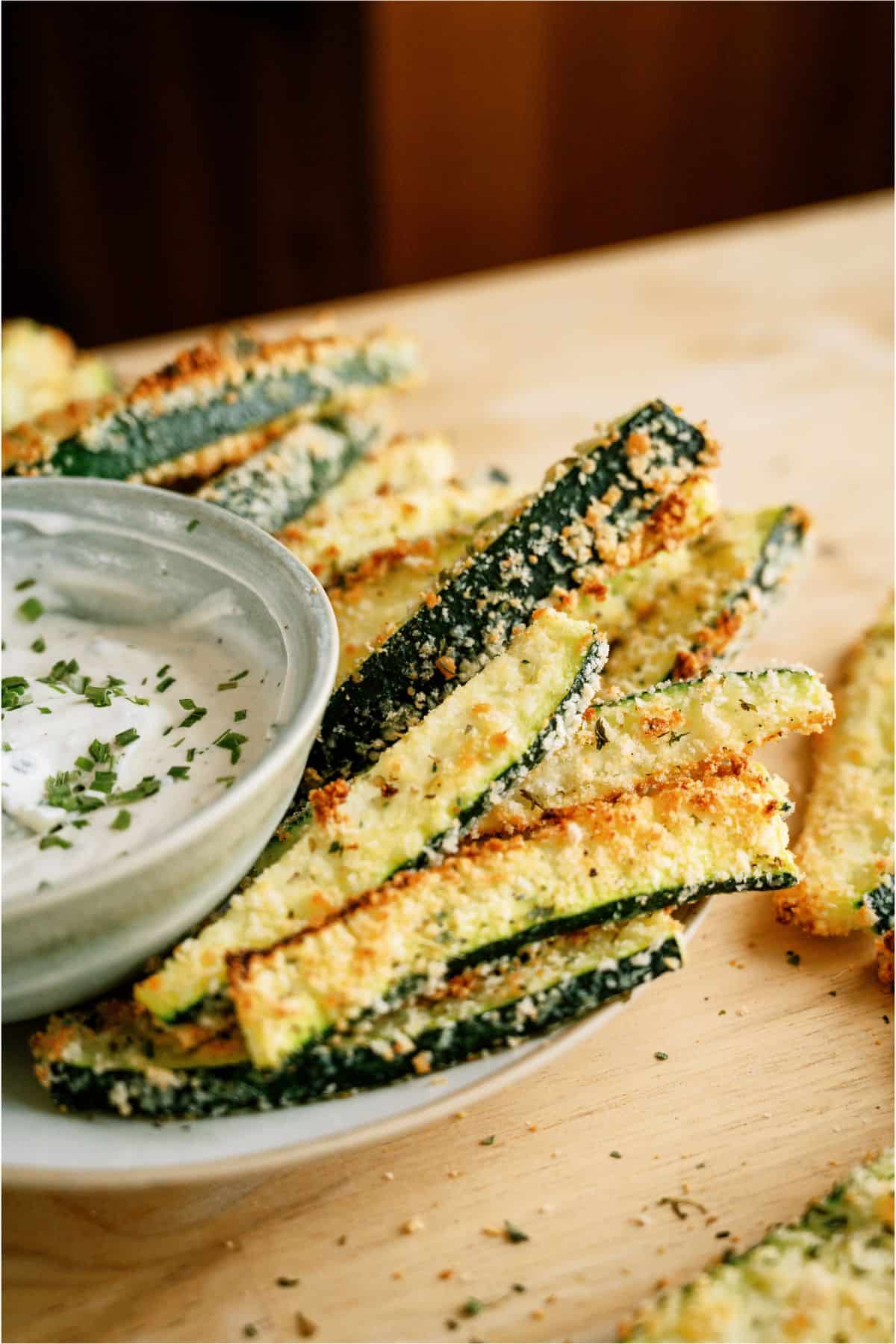 Plate of breaded zucchini sticks with a small bowl of dipping sauce, garnished with chopped herbs.