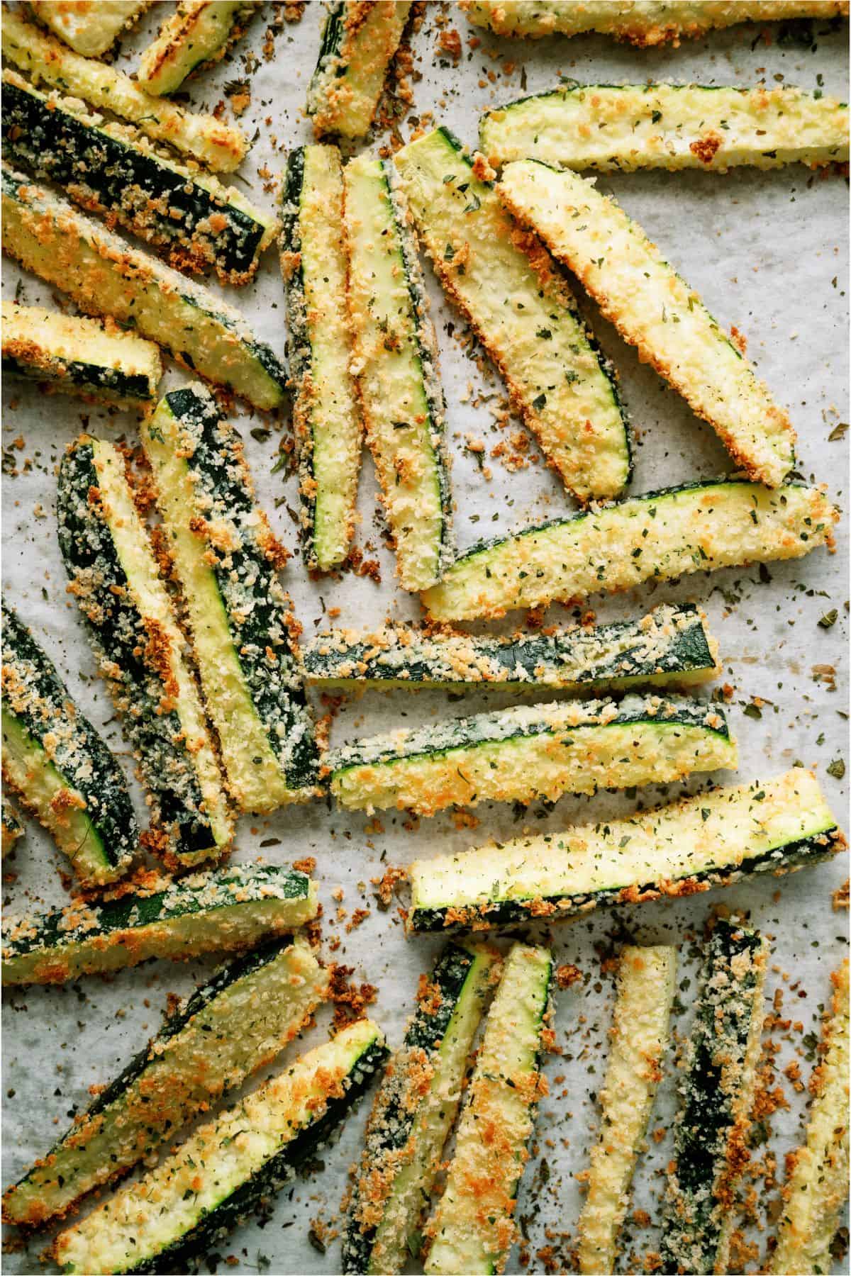 A plate of breaded zucchini fries is placed next to a bowl of creamy dip, garnished with chopped herbs, on a wooden surface.