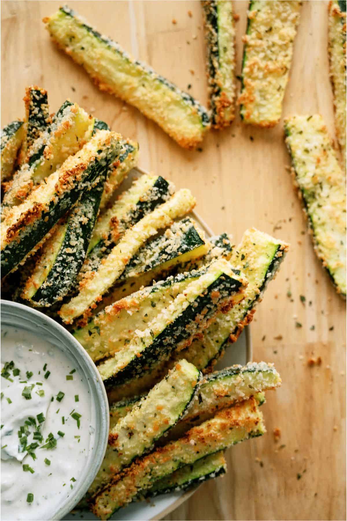 A plate of breaded zucchini fries is placed next to a bowl of creamy dip, garnished with chopped herbs, on a wooden surface.