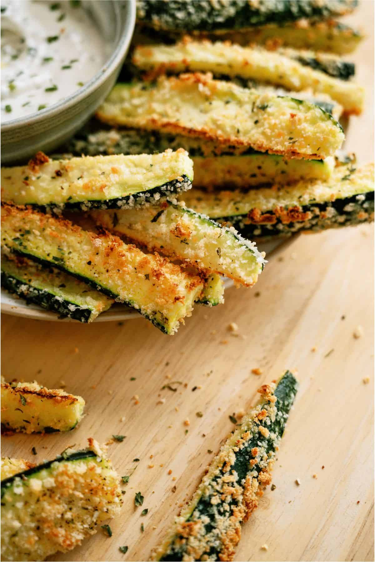 Close-up of baked zucchini fries coated with breadcrumbs and herbs on a wooden surface. A bowl of dipping sauce is in the background.