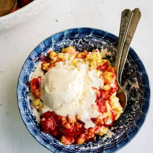 A blue patterned bowl filled with cherry cobbler topped with a scoop of melting vanilla ice cream, with two spoons placed inside.
