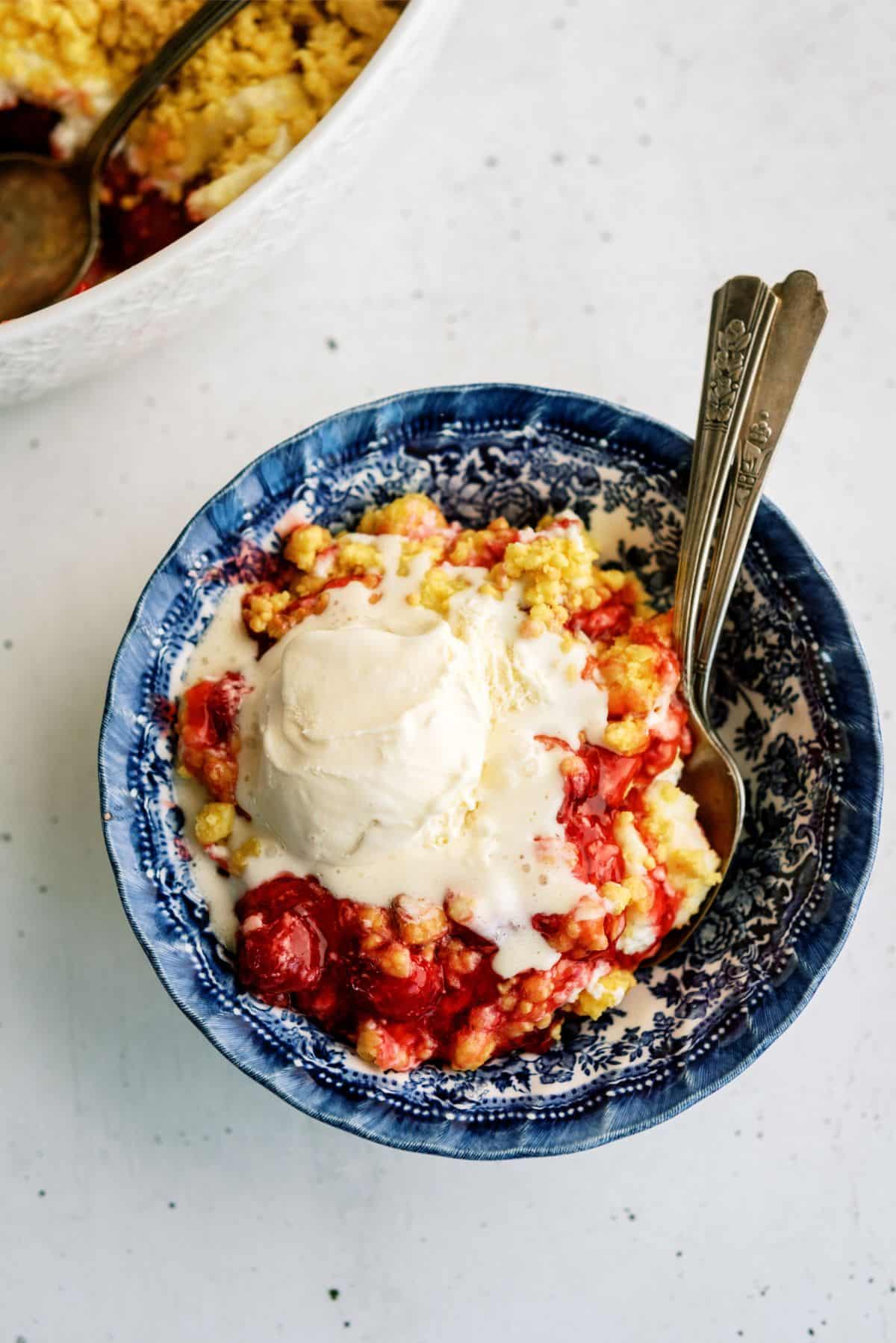 A blue patterned bowl filled with cherry cobbler topped with a scoop of melting vanilla ice cream, with two spoons placed inside.