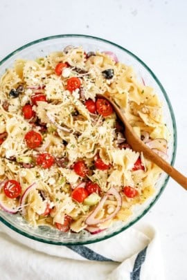 A glass bowl filled with pasta salad, featuring farfalle, cherry tomatoes, black olives, red onions, and grated cheese, with a wooden spoon resting inside.