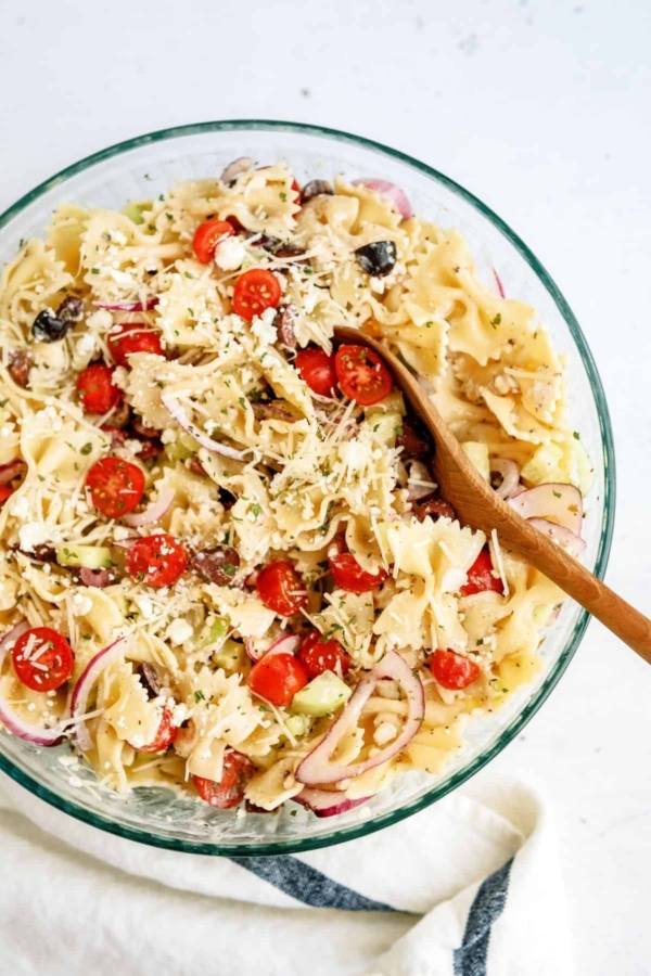 A glass bowl filled with pasta salad, featuring farfalle, cherry tomatoes, black olives, red onions, and grated cheese, with a wooden spoon resting inside.