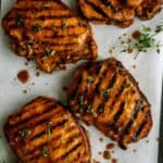 Four grilled pork chops with grill marks, garnished with fresh herbs and drizzled with sauce, arranged on parchment paper.