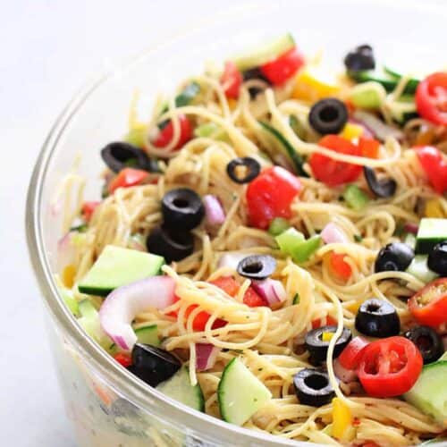 A clear bowl filled with pasta salad containing spaghetti, sliced cucumbers, cherry tomatoes, black olives, red onion, and bell peppers.
