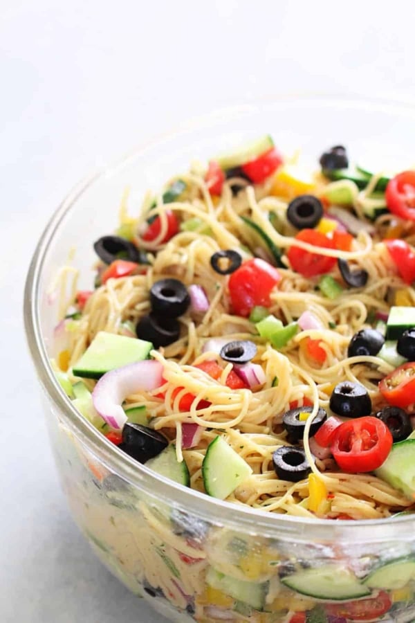 A clear bowl filled with pasta salad containing spaghetti, sliced cucumbers, cherry tomatoes, black olives, red onion, and bell peppers.