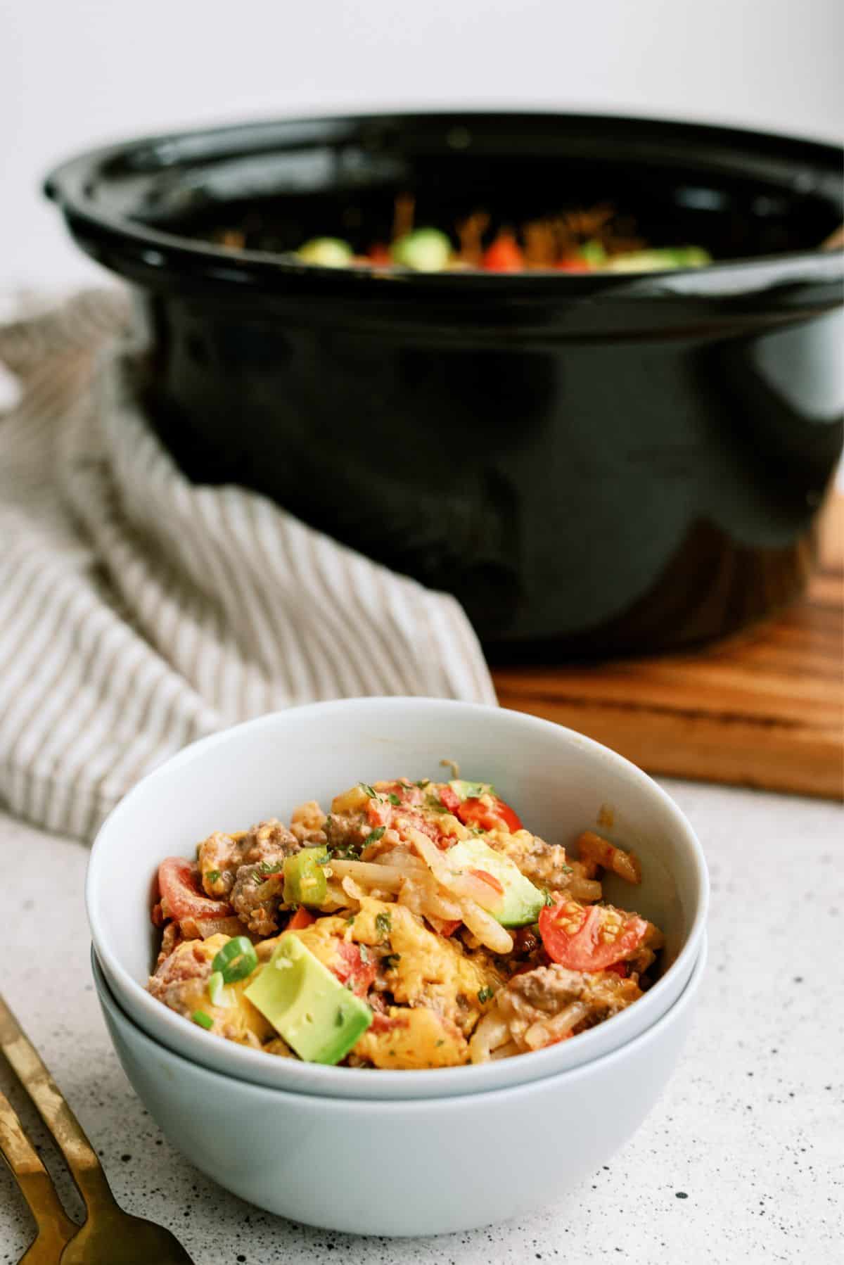 One serving of Slow Cooker Mexican Hamburger Hashbrown Casserole in a bowl with the slow cooker in the back ground