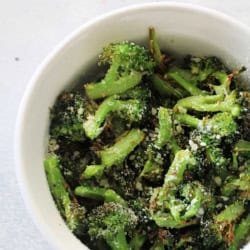 A white bowl filled with roasted broccoli florets topped with grated cheese, set on a light-colored surface.