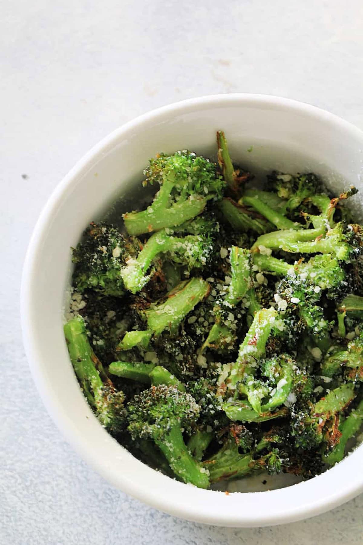 A white bowl filled with roasted broccoli florets topped with grated cheese, set on a light-colored surface.