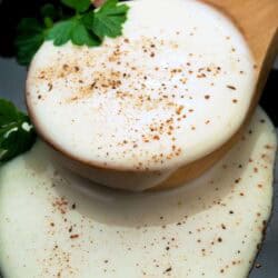 A wooden spoon with creamy white béchamel sauce sprinkled with ground pepper, garnished with parsley, resting on a dark surface.