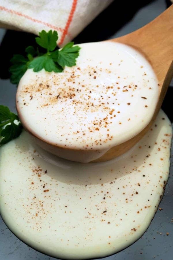 A wooden spoon with creamy white béchamel sauce sprinkled with ground pepper, garnished with parsley, resting on a dark surface.