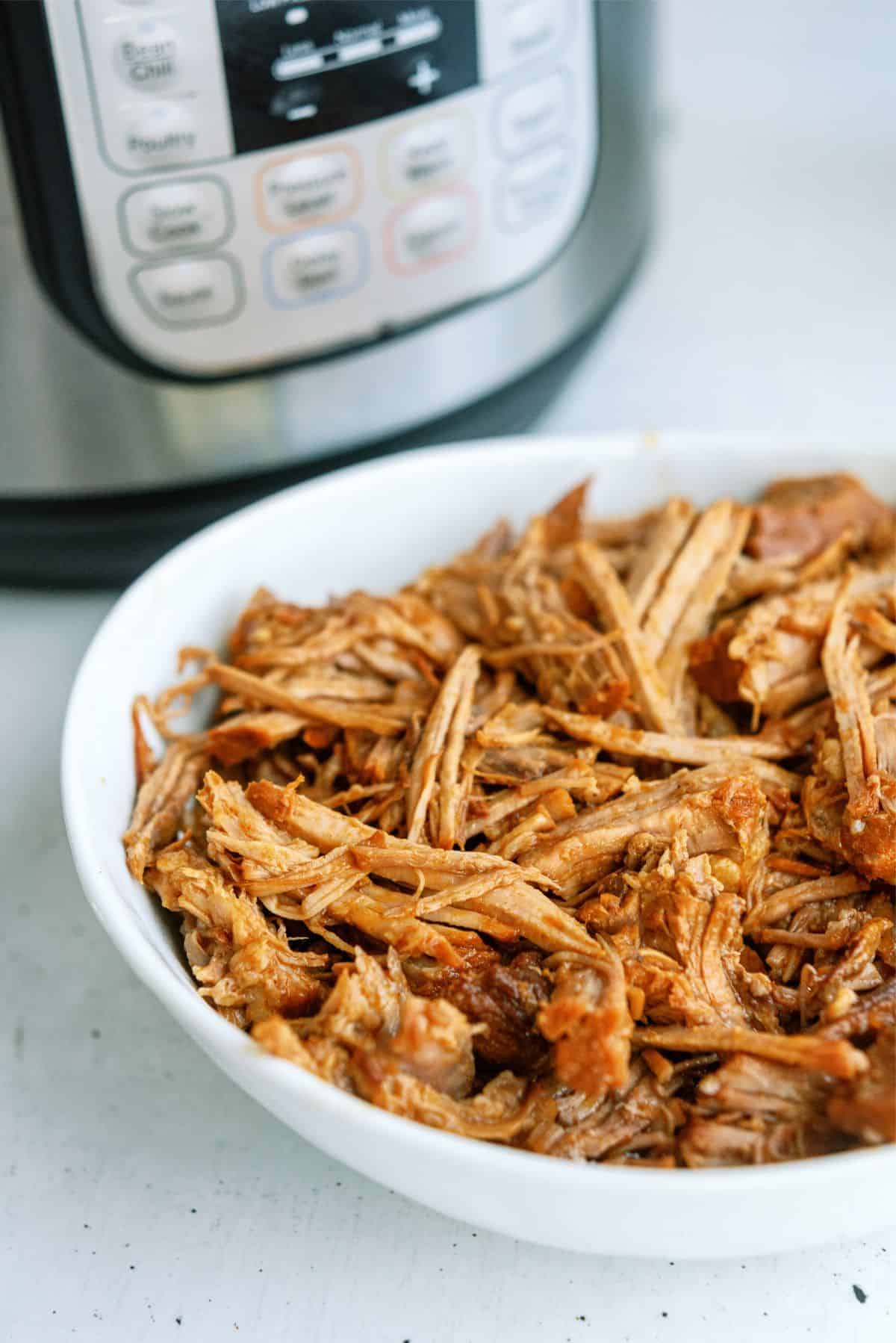 A white bowl filled with shredded cooked meat sits in front of an electric pressure cooker on a white surface.