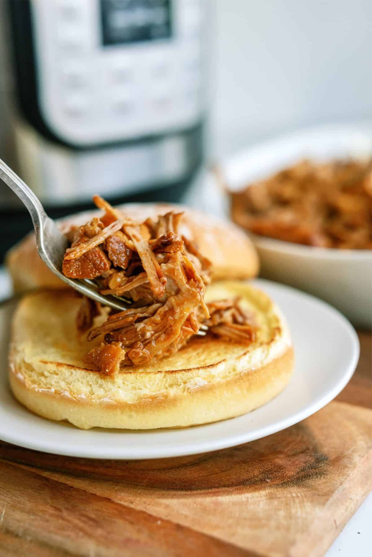 A fork places shredded barbecue meat onto a toasted sandwich bun on a white plate, with a bowl of meat and a kitchen appliance in the background.