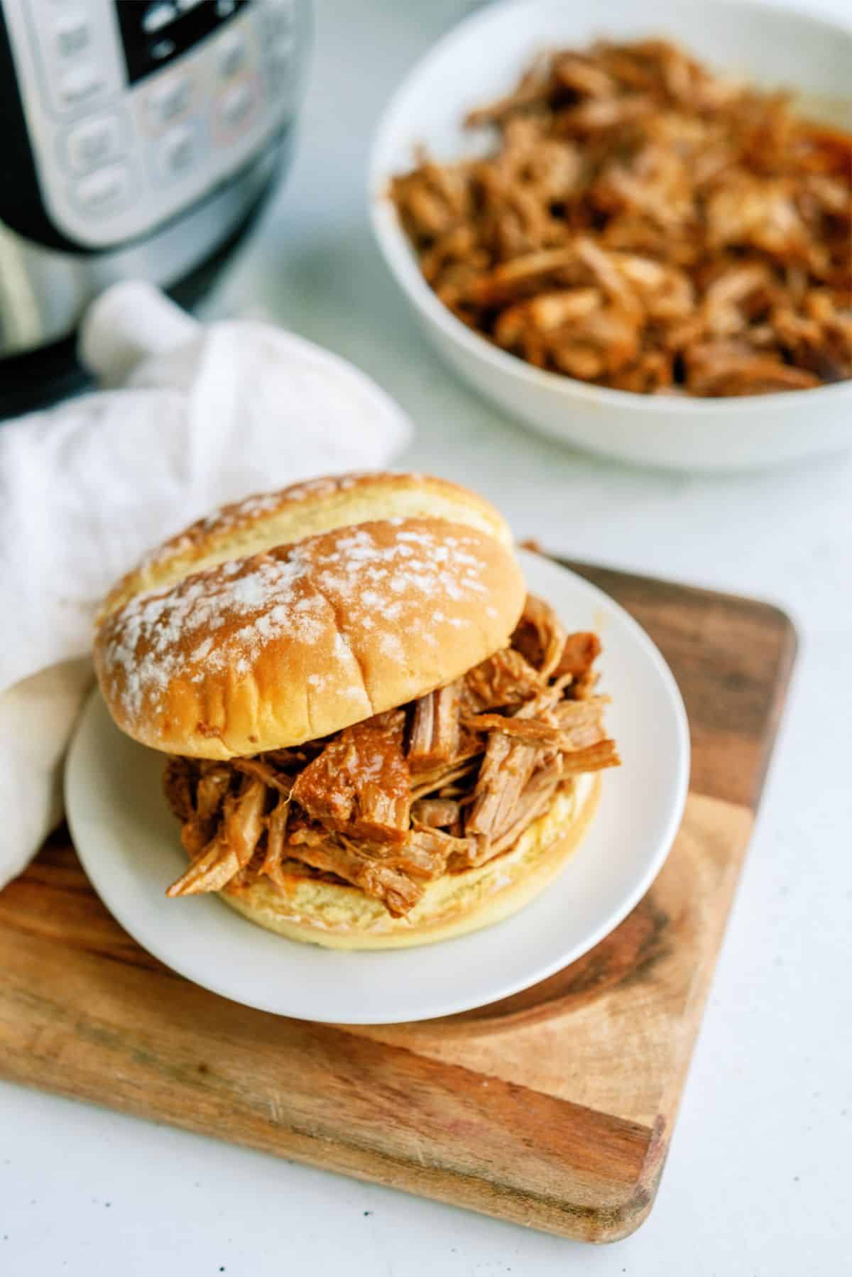 A pulled pork sandwich sits on a white plate atop a wooden board, with a bowl of pulled pork and a pressure cooker in the background.