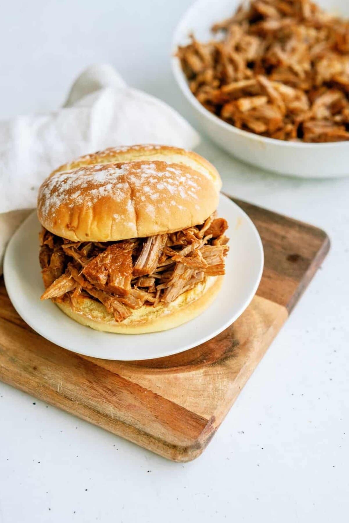 A pulled pork sandwich on a white plate sits on a wooden board, with a bowl of extra pulled pork in the background.
