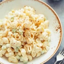 A bowl of creamy macaroni salad with chopped celery, topped with a sprinkle of seasoning, served on a rustic plate next to a fork and knife.