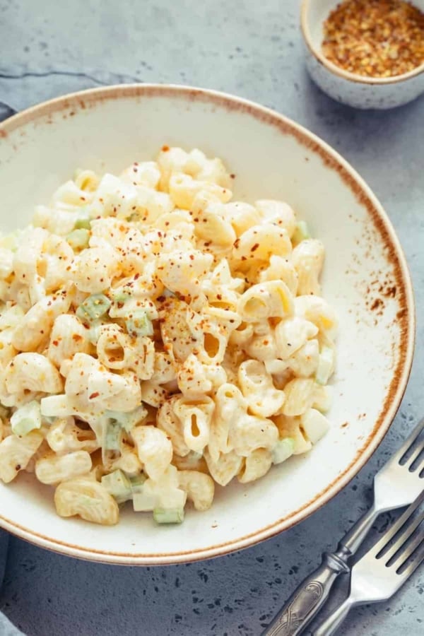 A bowl of creamy macaroni salad with chopped celery, topped with a sprinkle of seasoning, served on a rustic plate next to a fork and knife.