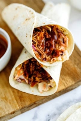 Close-up of two BBQ chicken wraps with shredded chicken, coleslaw, and barbecue sauce on a wooden surface. A small bowl of additional barbecue sauce is partially visible to the side.