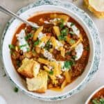 A bowl of chili topped with shredded cheese, sour cream, and chopped herbs, served with buttered cornbread on the side and a spoon in the bowl.