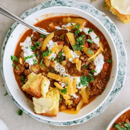 A bowl of chili topped with shredded cheese, sour cream, and chopped herbs, served with buttered cornbread on the side and a spoon in the bowl.