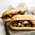 Two beef sandwiches with melted cheese and sautéed vegetables on a cutting board.