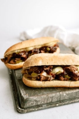 Two beef sandwiches with melted cheese and sautéed vegetables on a cutting board.