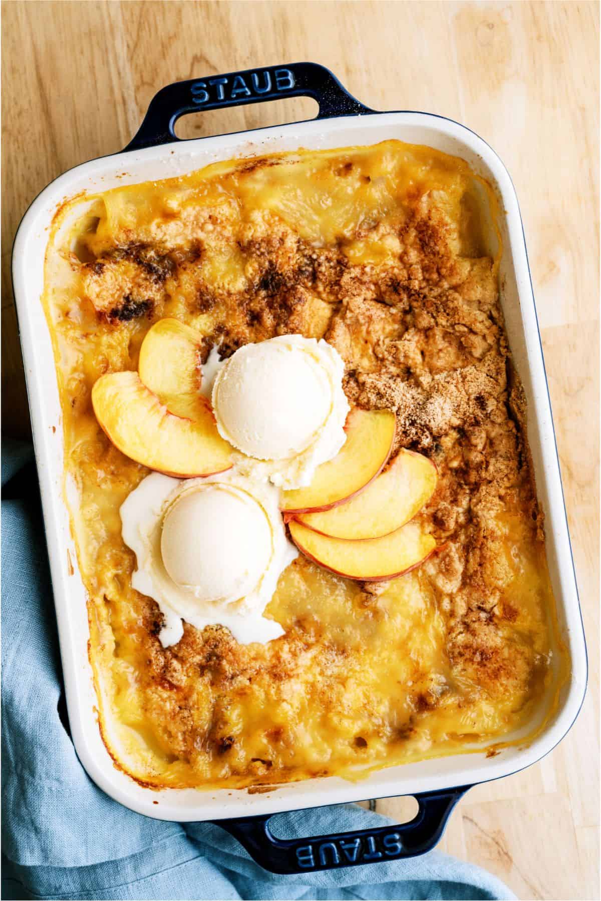 Baked peach cobbler in a rectangular dish, topped with two scoops of vanilla ice cream and fresh peach slices.