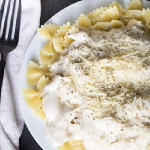 A plate of bowtie pasta topped with a creamy white sauce and grated cheese, with a fork on a napkin beside the plate.