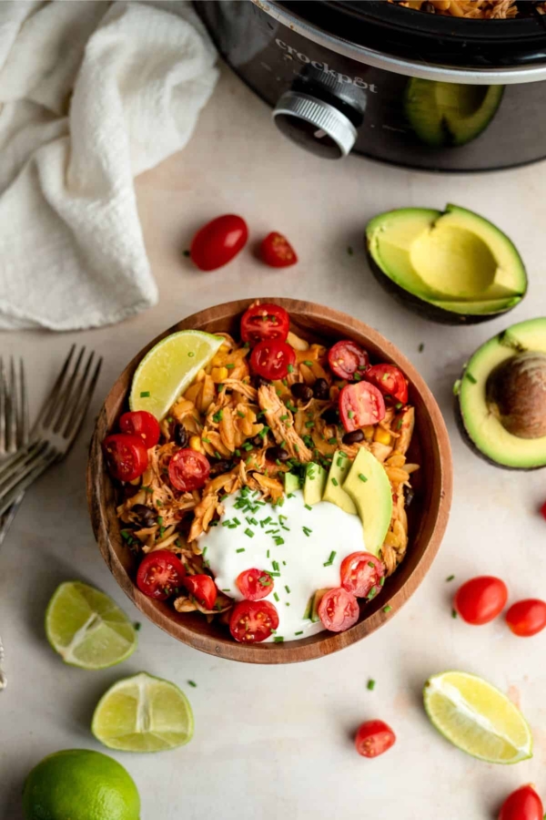 A wooden bowl filled with shredded chicken, avocado slices, cherry tomatoes, lime wedge, and a dollop of sour cream, garnished with chopped herbs. Forks, avocados, tomatoes, and lime pieces surround it.