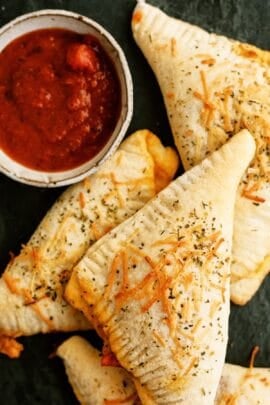 Three baked pizza pockets topped with herbs and cheese are arranged next to a small bowl of marinara sauce for dipping.