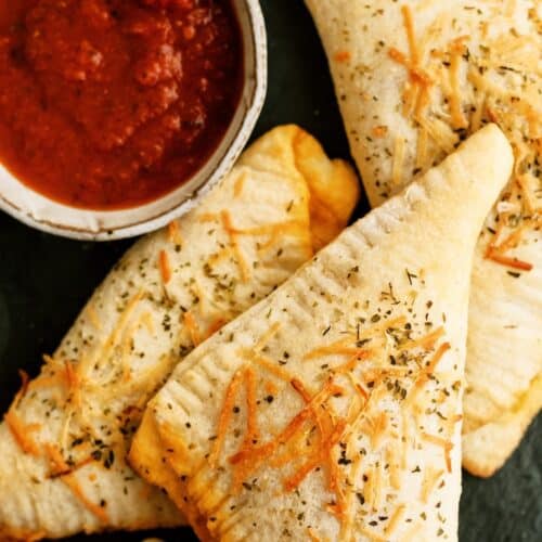 Three baked pizza pockets topped with herbs and cheese are arranged next to a small bowl of marinara sauce for dipping.