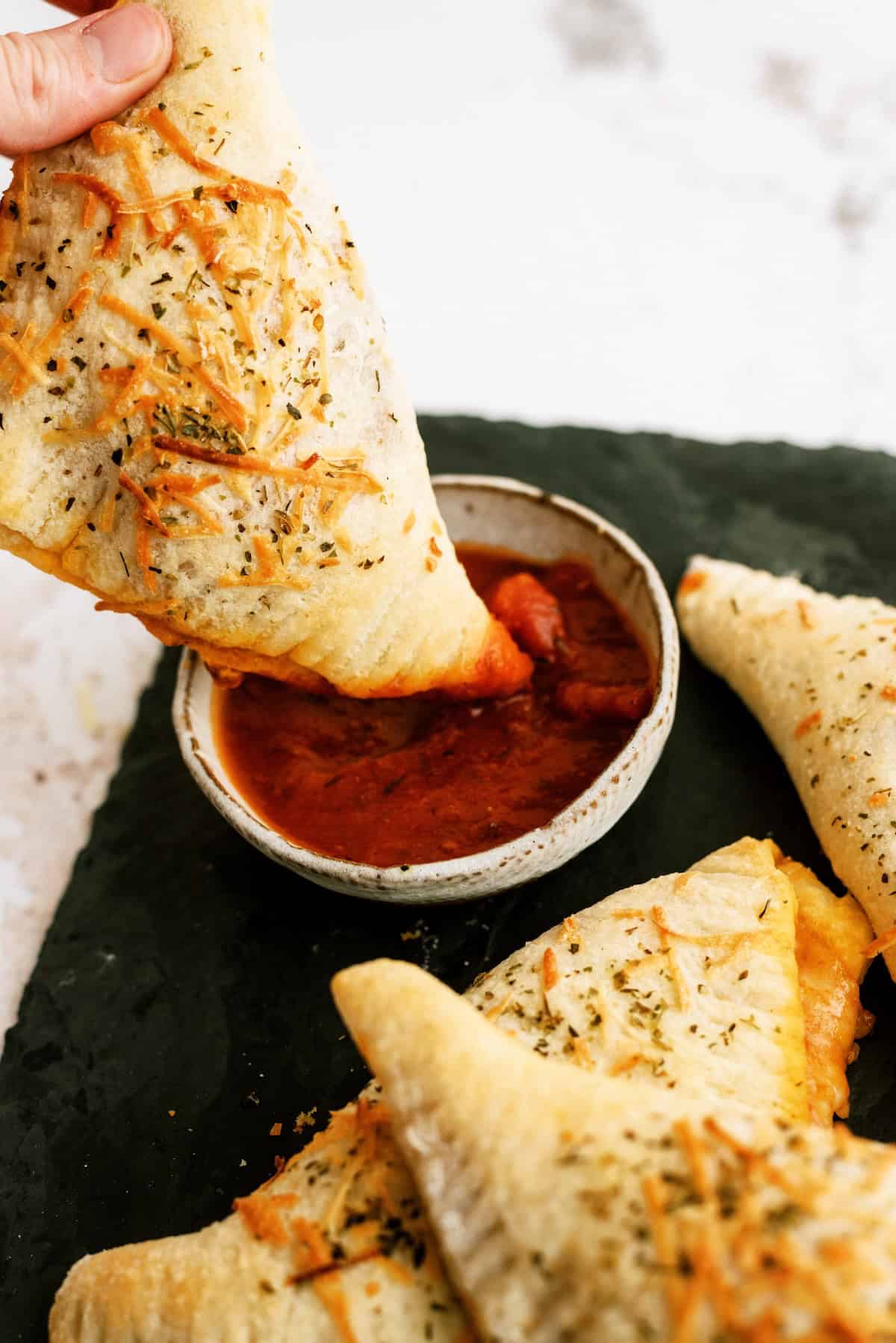 A hand dips a baked calzone topped with herbs and cheese into a bowl of marinara sauce, with other calzones on a dark surface nearby.