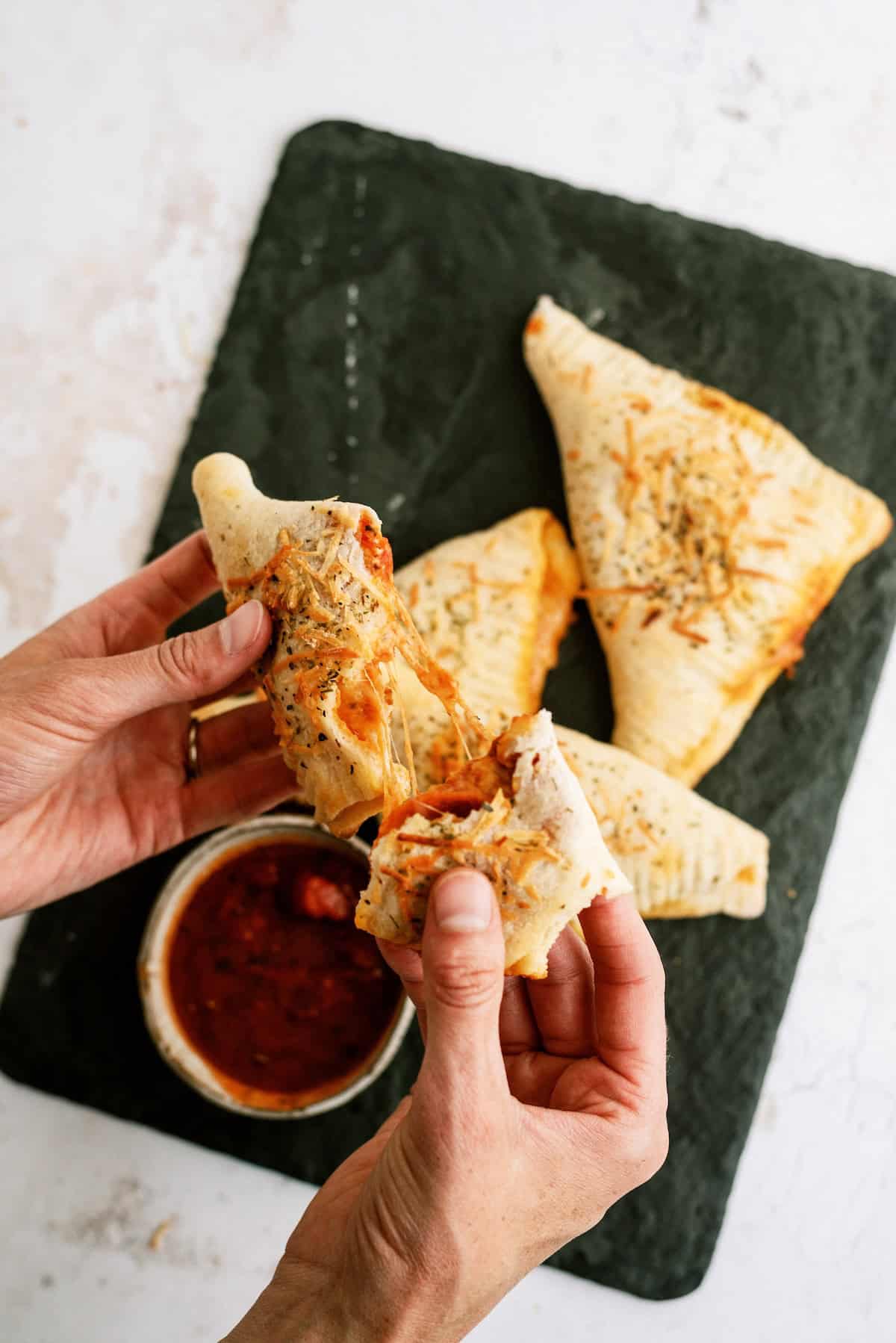 A person pulls apart a baked pizza pocket with melted cheese, with more pizza pockets and a bowl of marinara sauce on a black slate.