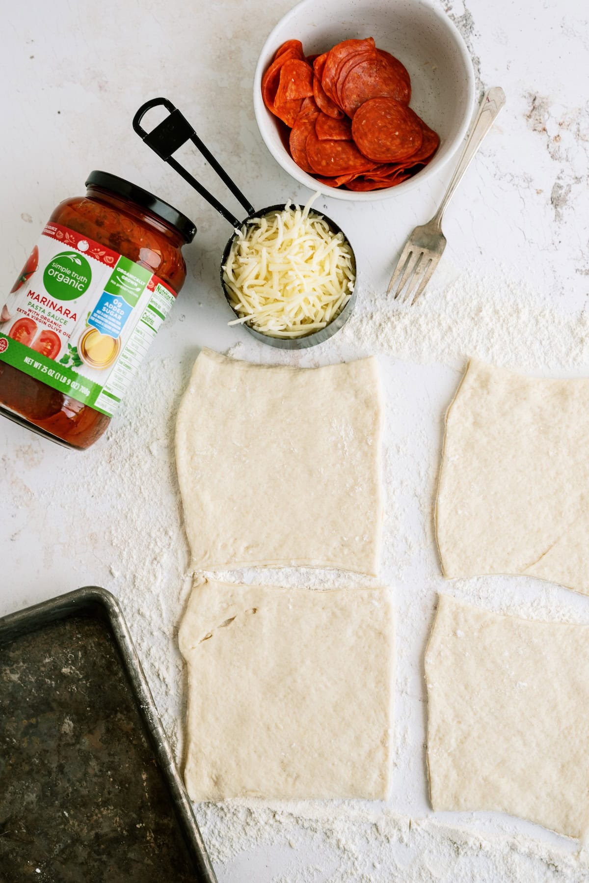 Four rectangular pieces of pizza dough on a floured surface, with a jar of marinara sauce, a bowl of pepperoni, shredded cheese, a fork, and a baking tray nearby.