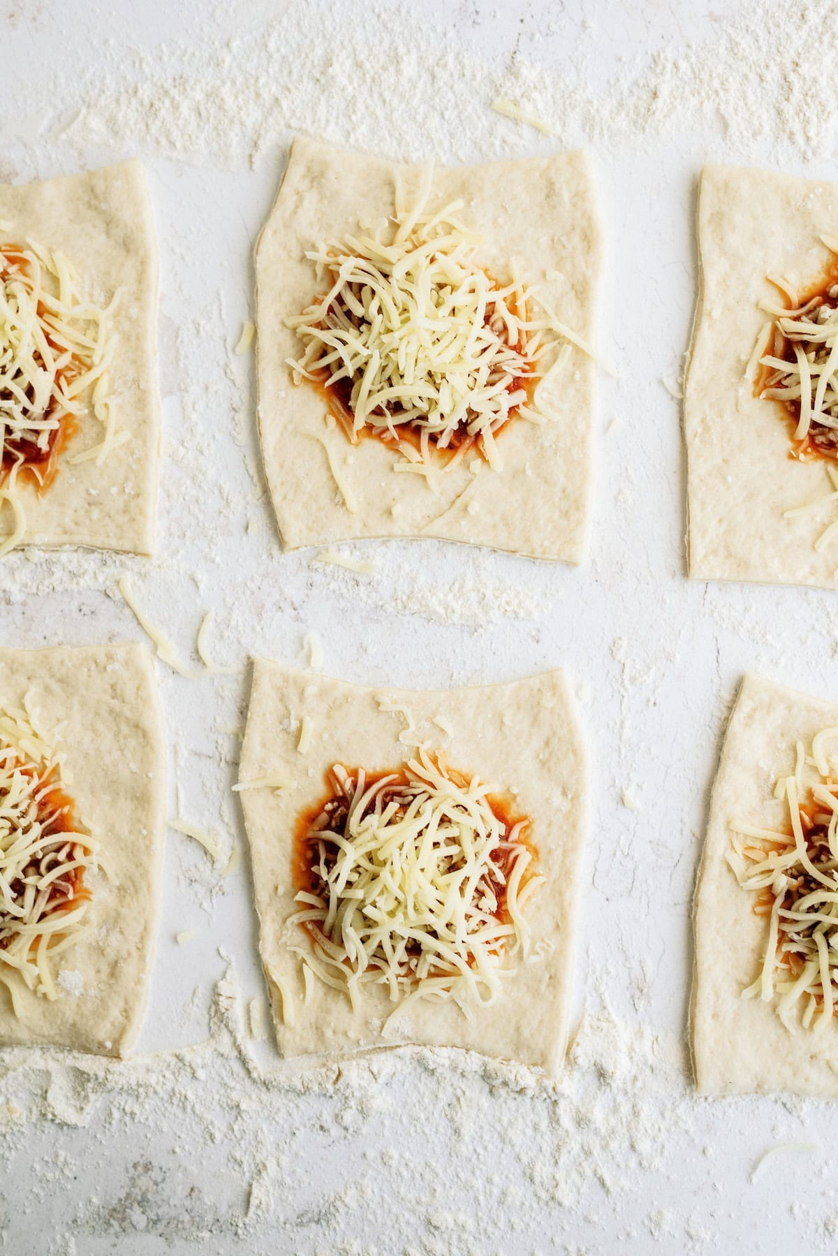 Six square pieces of dough topped with tomato sauce and shredded cheese are arranged on a floured surface, ready to be baked or folded.