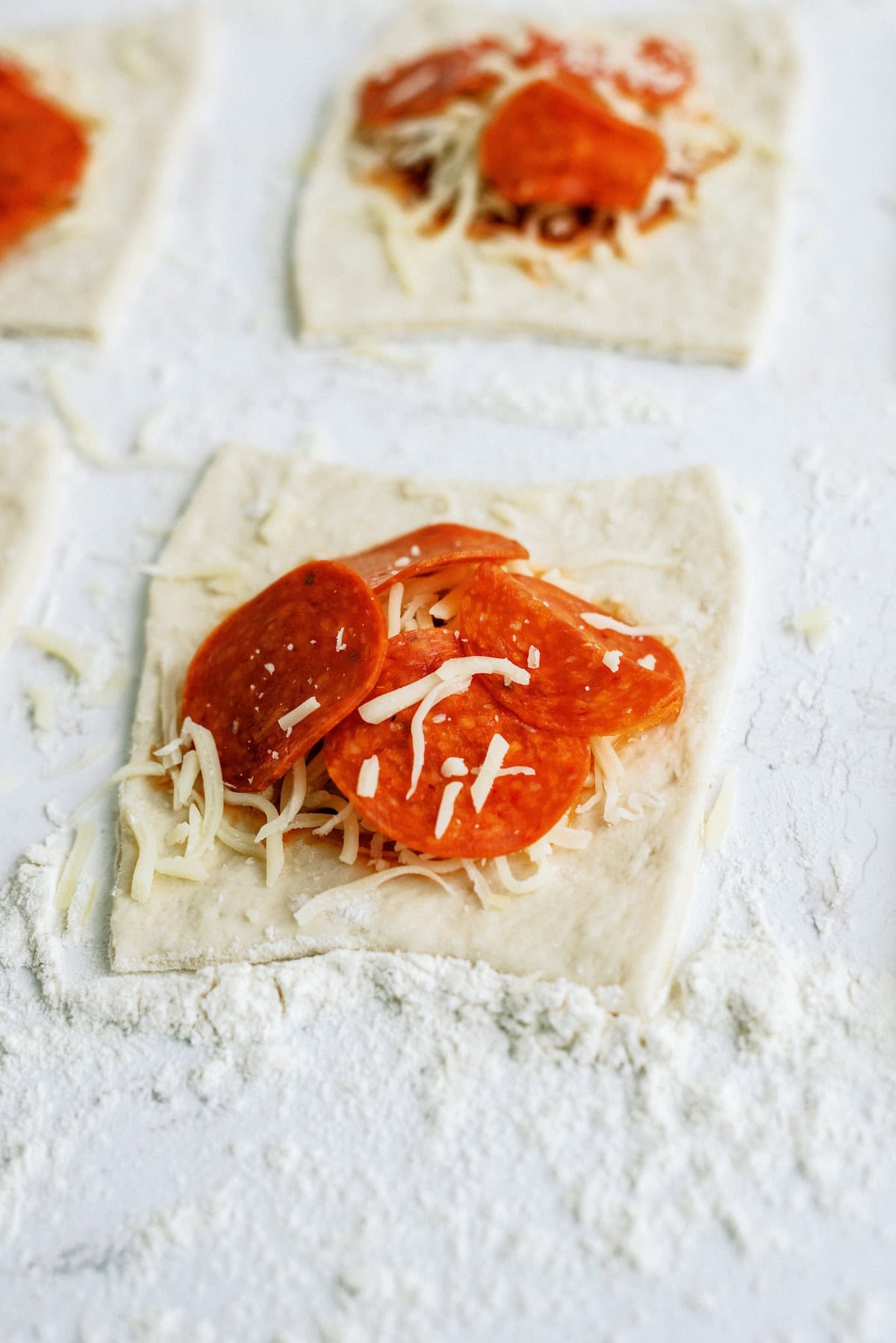 Rectangles of dough topped with shredded cheese and pepperoni slices are arranged on a floured surface, ready to be folded or baked.