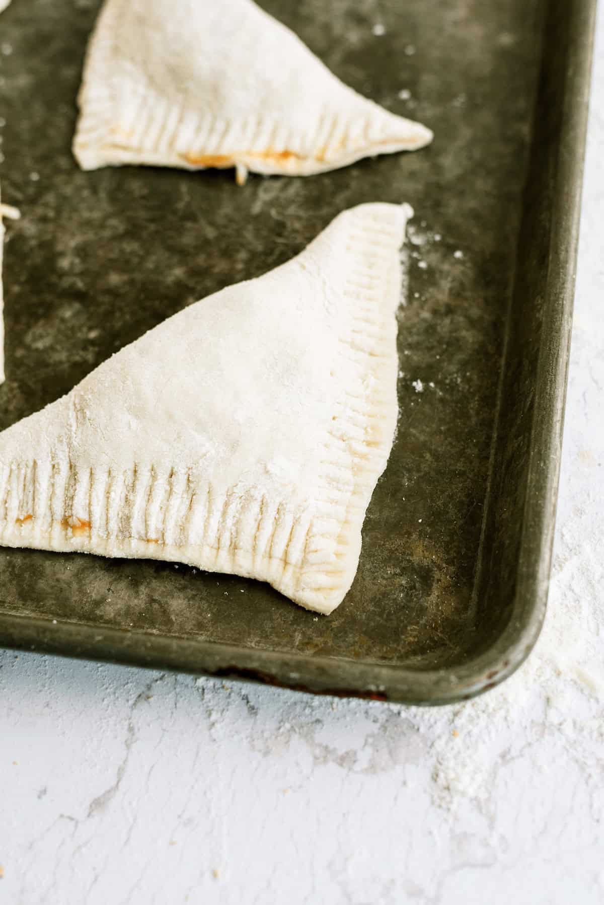 A raw, triangular pastry with crimped edges sits on a floured baking sheet, ready to be baked.